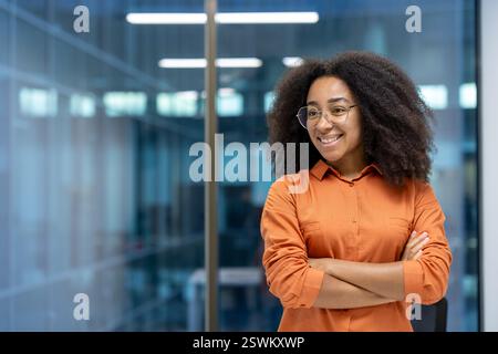 Young adult African American woman stands confidently with crossed arms in a modern office setting, smiling and looking away Stock Photo