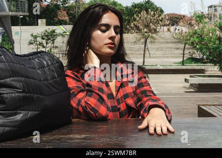 Young woman wearing a red and black checkered shirt is sitting at a wooden table in a park, touching her neck with a thoughtful expression Stock Photo