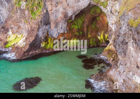 A large rock formation with a small cave in it. The cave is green and has a lot of moss growing on it. The water is blue and there are some rocks in t Stock Photo