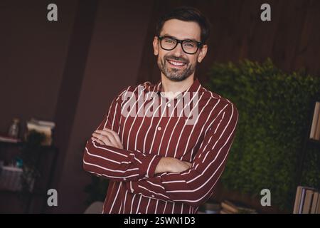 Young bearded man wearing striped shirt smiling indoors, showing a relaxed and confident posture in a stylish and comfortable interior. Stock Photo