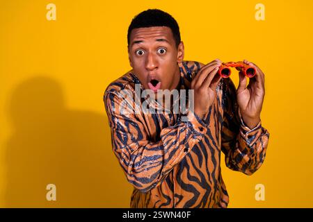 Young man in stylish shirt looks surprised holding red binoculars against yellow background Stock Photo