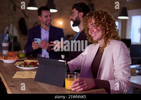 Businesswoman using tablet and drinking juice during office break with colleagues chatting Stock Photo