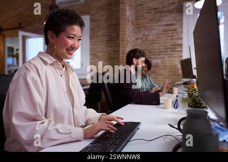 Young asian woman working on computer in busy office with colleagues collaborating Stock Photo