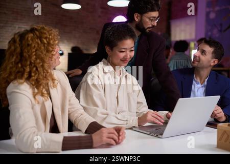 Diverse business team collaborating on laptop in modern office at night Stock Photo