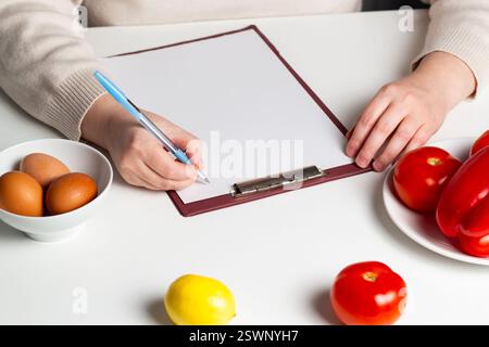 Blank mock up of diet plan, diet menu. Paper with copy space on table with vegetables and eggs. Woman preparing healthy eating plan. Stock Photo
