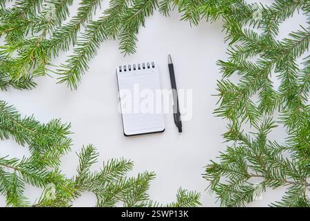 Blank spiral notepad and black pen placed on white surface, framed by fresh green fir branches. Minimalistic and nature-inspired composition, ideal fo Stock Photo