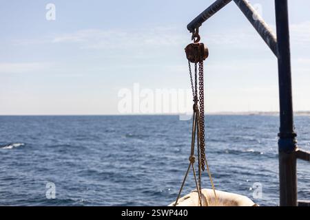 Rusty pulley and chains hanging from a metal jib over a calm blue sea, creating a serene maritime scene in marsa alam, egypt Stock Photo