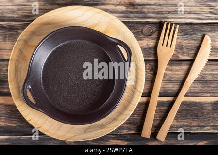Empty black ceramic plate in a pan style on a wooden surface, next to some cutlery, on a dark rustic wooden table. Stock Photo