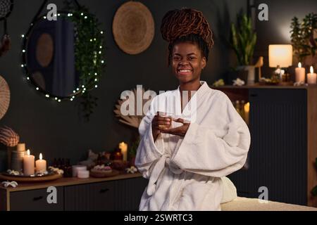 Medium close up of African American young pretty woman in bathrobe holding transparent cup of herbal tea smiling while posing for portrait Stock Photo