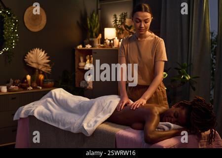 Beautiful young woman lying on table and enjoying adorable massage with essential oils by Caucasian masseuse Stock Photo