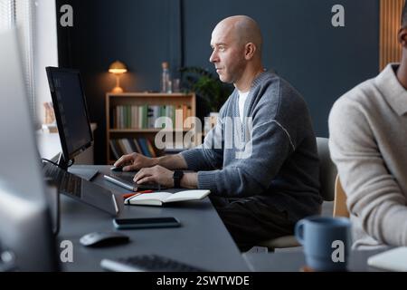 Side view of concentrated mature man with shaved head using desktop computer at working station against dark blue wall in corporate office Stock Photo
