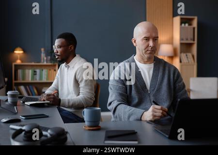 Medium shot of middle aged male interior designer with shaved head drawing on pen tablet concentrating on laptop screen, while working at desk with colleague in background in office Stock Photo