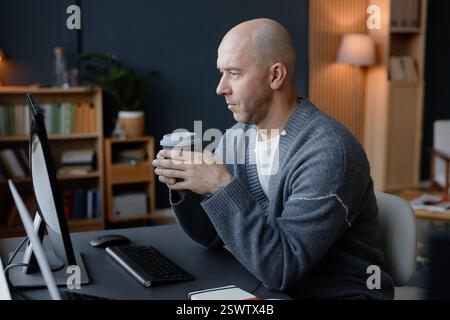Side view of thoughtful mature man with shaved head holding reusable cup in hands looking at computer screen, while planning work at desk in corporate office Stock Photo