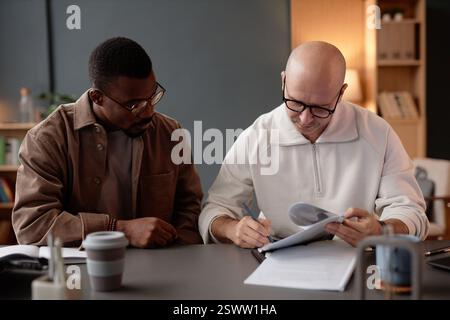 Medium shot of mature man with shaved head focused on signing document working with Black male employee sitting at desk in modern office setting Stock Photo