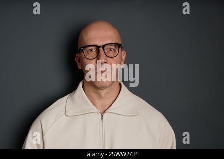 Studio portrait of confident middle aged man with shaved head wearing casual sweatshirt and glasses posing against dark grey wall, camera flash, copy space Stock Photo