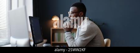 Website header shot of thoughtful Black office worker using desktop computer at working desk against dark blue wall in home office, copy space Stock Photo