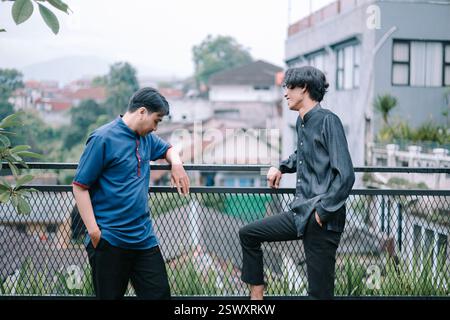 Two young men in stylish casual attire stand on an urban rooftop balcony, engaged in a pleasant conversation. Their relaxed postures and the city back Stock Photo