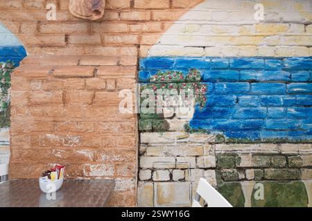 Painted brick wall, likely in Venice, Italy, featuring a mural of a tree and blue water.  Outdoor cafe setting with condiments on a table. Stock Photo