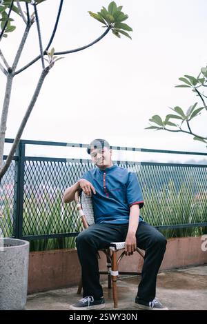 A young man in casual attire sitting comfortably on a chair on a rooftop terrace. He is smiling gently and appears relaxed, enjoying a calm moment out Stock Photo