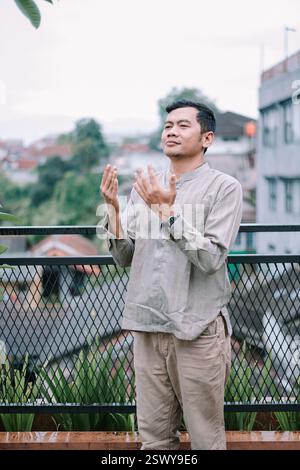 A young muslim man praying with raised hands on a balcony. He is experiencing spiritual gratitude and devotion in a cityscape setting. Demonstrating f Stock Photo