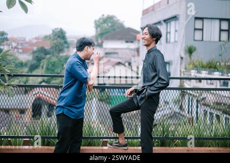 Two young Asian men laugh and chat on a rooftop terrace. They are dressed in casual shirts and appear to be enjoying a lighthearted moment of friendsh Stock Photo