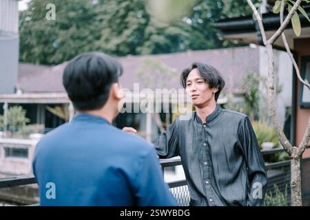 Two young men engage in a casual and friendly conversation outdoors on a leafy balcony. They are relaxing, enjoying each other's company, and sharing Stock Photo