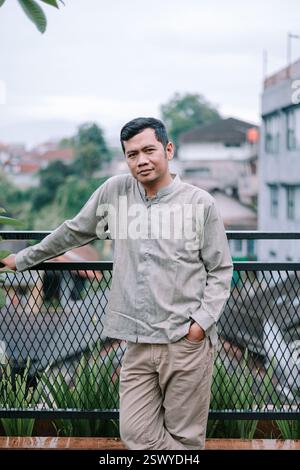 A thoughtful Asian man stands on a terrace, leaning against the railing, gazing directly at the camera. He is dressed casually in khakis and a pattern Stock Photo