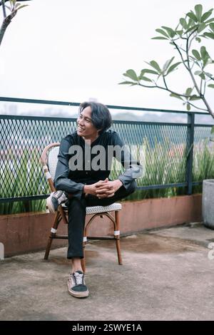 An asian man is sitting on a wicker chair on an outdoor terrace overlooking a cityscape. He is looking to the distance with a pensive and thoughtful e Stock Photo