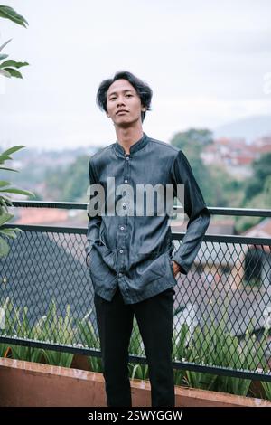 A young Asian man dressed in a casual shirt and black pants is posing outside with his hands in his pockets. The background reveals a hillside town cr Stock Photo