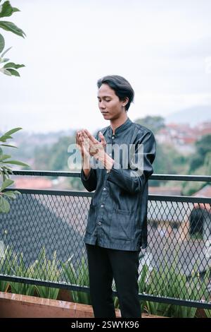 A young man is seen in a moment of serene prayer on a balcony, gazing downwards with hands raised in devotion. The cityscape fades gently into the bac Stock Photo
