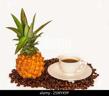Exotic Mini Pineapple and Elegant White Coffee Cup on a Saucer, Placed on a Bed of Roasted Coffee Beans Against a Clean White Background Stock Photo