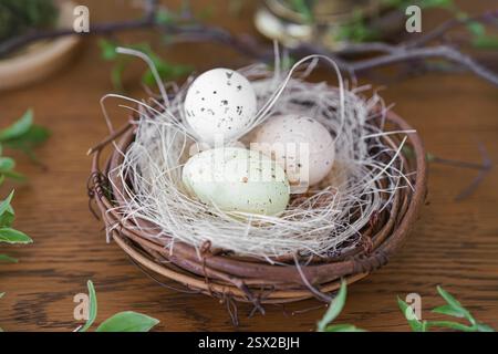 Three colorful eggs in natural straw nest surrounded by green leaves on brown wooden surface. Easter holiday home decor, spring background Stock Photo