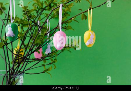 Colorful Easter eggs with cute white bunnies hanging on green tree branches in glass vase, green background. Easter concept. Easter still life. Stock Photo