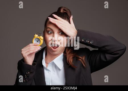 Stressed businesswoman in a suit holding a small alarm clock, shocked at the time, running late, concept of time management and deadline pressure Stock Photo