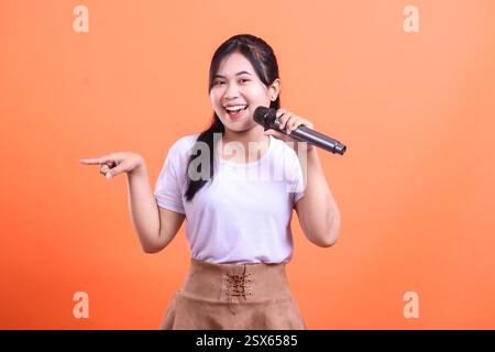 A young woman singing with a microphone and pointing with her left hand in a studio, she looks very happy and friendly. isolated on orange background Stock Photo