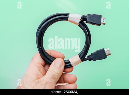 A person holds a neatly coiled black HDMI cable with two connectors, secured by paper tape, against a clean green background in a well-lit setting. Stock Photo
