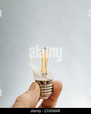 A person holds a shattered LED filament bulb, exposing internal yellow filaments and wiring against a neutral background, highlighting the fragile str Stock Photo