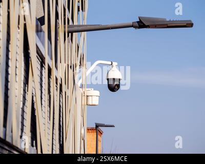 A white dome-shaped security camera with a black lens is attached to a metal arm on the side of a modern building, monitoring the area against a clear Stock Photo