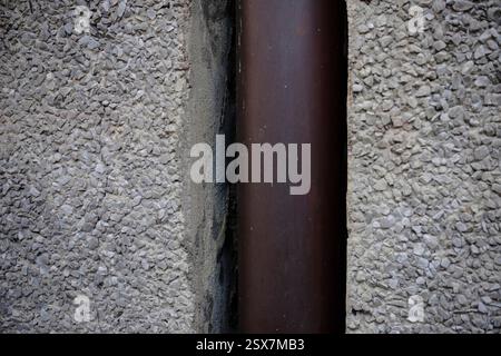 Cremona, Italy February 9th 2025 Vertical close up of a brown downspout crossing a concrete wall featuring a coarse gravel finish, creating a contrast Stock Photo