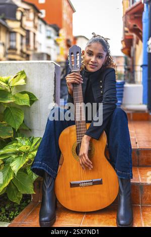 Vertical portrait of a young african american woman with her guitar sitting outside a colorful patio of a house in the city Stock Photo