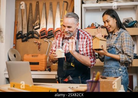 Senior carpenter and woman in a workshop presenting model houses to customers on a laptop via live Stock Photo