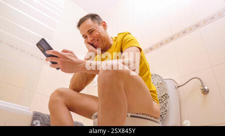 Man sitting on toilet using smartphone, laughing and enjoying Stock Photo