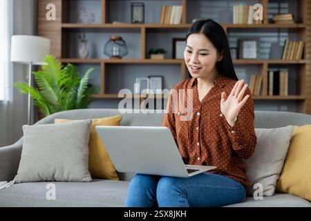 Asian woman using laptop for video call communication at home. Smiling and waving while connecting online. Cozy setting with sofa and shelves. Emphasis on digital interaction and comfort. Stock Photo