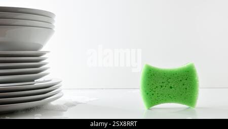Conceptual detail of hand washing dishes with green sponge scouring pad and stack of white plates on white bench with water and foam and isolated back Stock Photo