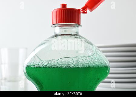 Detail of top of plastic bottle with dishwasher product with open cap and glass plates and glasses in the background isolated on white background. Fro Stock Photo