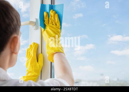 Man in yellow gloves wipes a window with sponge cloth, letting in spring sunlight and fresh air and revealing blue sky Stock Photo