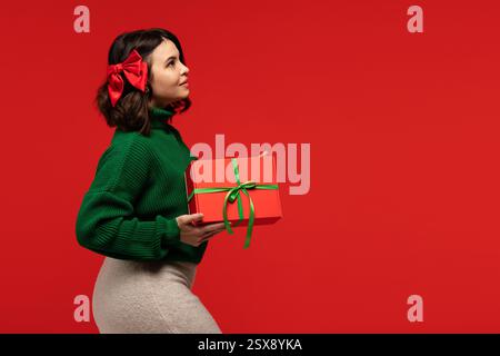 Young woman in a green sweater with a red bow in her hair, holding a wrapped red gift box against a red background. Stock Photo