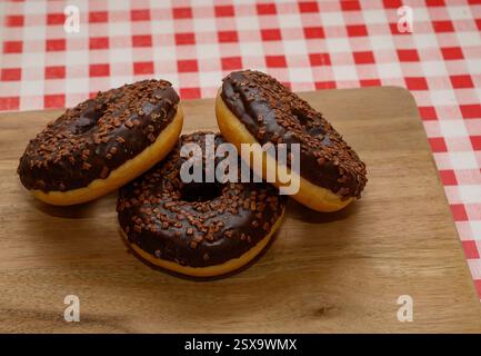 Three chocolate glazed donuts topped with sprinkles are arranged neatly on a wooden board. The background features a classic red and white checkered t Stock Photo