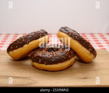 Three chocolate glazed donuts topped with sprinkles are arranged neatly on a wooden board. The background features a classic red and white checkered t Stock Photo
