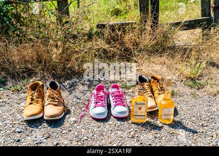 Two bottles, Teacher's Scotch whiskey and the Kings Ginger Liqueur, with three pairs of trainers and walking shoes on side of road by a field. Stock Photo